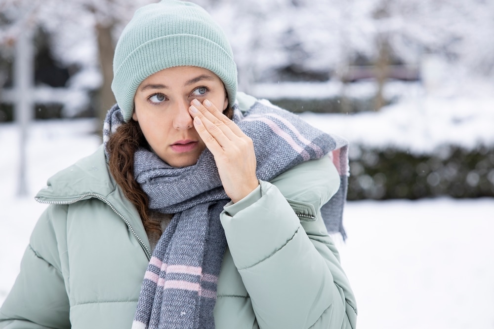 Young woman, wearing winter clothing, rubs her dry eyes during snowy weather